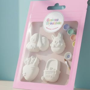 Può includere: Una confezione rosa contenente quattro statuette pasquali in gesso bianco: un coniglietto, un pulcino, una carota e un cestino con la scritta "Happy Easter". Include vasetti di vernice e un pennello. La confezione ha un adesivo "Coucou Creatives".
