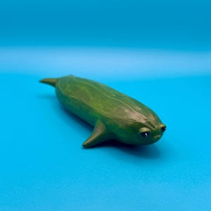 May include: A small, smooth, green figurine shaped like a cucumber with a tapered tail, two side fins, and two small, dark, glossy eyes positioned on the front. The object has a textured, organic surface with subtle ridges and a rounded body.