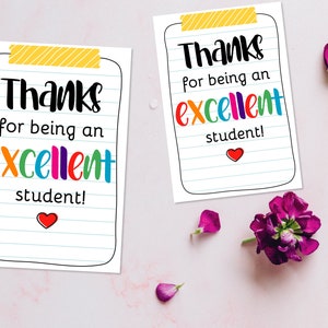 K&ouml;nnte beinhalten: Zwei wei&szlig;e Karten mit dem Text "Danke, dass du ein ausgezeichneter Sch&uuml;ler bist!" in schwarzen und bunten Buchstaben. Die Karten sind wie Notizpapier gestaltet, mit gelbem Klebeband oben und einem roten Herz unten. Eine lila Blume und Bl&uuml;tenbl&auml;tter im Hintergrund.