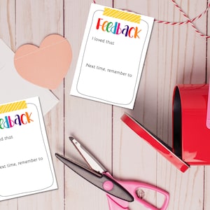 Könnte beinhalten: Zwei weiße Karten mit dem Wort "Feedback" in Regenbogenfarben und leeren Zeilen zum Schreiben. Die Karten liegen auf einer hellbraunen Holzoberfläche mit einer roten Briefkast, einem rosa Herzen und einer pinkfarbenen Schere.
