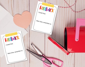 May include: Two white cards with the word "Feedback" in rainbow letters and blank lines for writing. The cards are on a light wood surface with a red mailbox, a pink heart, and a pair of pink and black scissors.