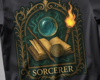 Op de afbeelding: Zwart T-shirt met een gedetailleerde illustratie van de gereedschappen van een tovenaar. Het ontwerp omvat een open boek, een gloeiende kristallen bol, een toverstaf en een brandende fakkel, allemaal binnen een sierlijk kader. Het woord "SORCERER" staat onderaan.
