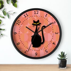 May include: A black and white cat clock with a pink and white geometric pattern background. The clock has black hands and black numbers.