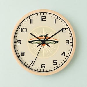 May include: A round wall clock with a light wood frame and a cream-colored face. The clock features black numerals from 1 to 12 and black hands. In the center, there is a graphic illustration of a flying saucer emitting light rays.