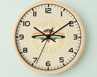 May include: A round wall clock with a light wood frame and a cream-colored face. The clock features black numerals from 1 to 12 and black hands. In the center, there is a graphic illustration of a flying saucer emitting light rays.