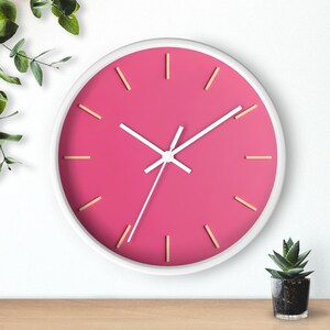May include: A round wall clock with a white frame and a vibrant pink face. The clock features white hands and gold-coloured hour markers. The clock is a decorative item for home decor.