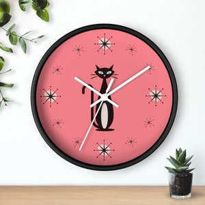 May include: A pink clock with black hands and a black cat silhouette in the center. The clock face has a mid-century modern design with white starbursts.