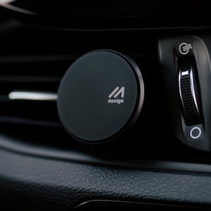 Könnte beinhalten: Schwarze Handyhalterung für das Auto mit dem Logo "M design", befestigt an einer Lüftungsöffnung im Auto. Das Armaturenbrett verfügt über einen Warnblinklichtschalter und Lüftungsregler. Die Halterung ist rund.