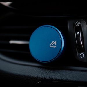 Może przedstawiać: Niebieski, okrągły uchwyt samochodowy na telefon z napisem "M design". Przymocowany do czarnej deski rozdzielczej samochodu. Wnętrze samochodu zawiera czerwony przycisk świateł awaryjnych. Około 5 cm średnicy.