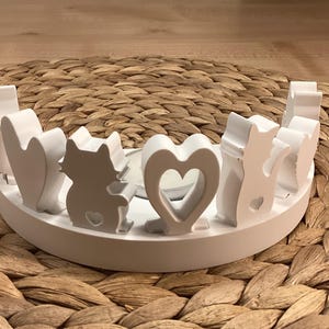 Könnte beinhalten: Ein weißer, halbkreisförmiger Kerzenhalter mit katzen- und herzförmigen Aussparungen. Der Halter hat ein niedriges Profil und besteht aus einem festen, undurchsichtigen Material. Das Design zeigt ein sich wiederholendes Muster aus Katzen und Herzen.