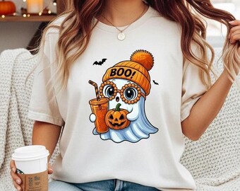 May include: Cream-colored t-shirt featuring a cartoon ghost wearing a knit hat with the word "BOO!" and glasses. The ghost is holding a pumpkin and a drink. Two black bats are flying above the ghost. The shirt is paired with blue jeans.