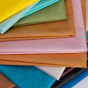 Puede incluir: Una colección de láminas de tela plegadas en varios colores, incluyendo amarillo, turquesa, verde oliva, bronceado, rosa claro, marrón, crema, azul oscuro, negro y rojo intenso. Cada pieza de material presenta un acabado superficial texturizado y arrugado.