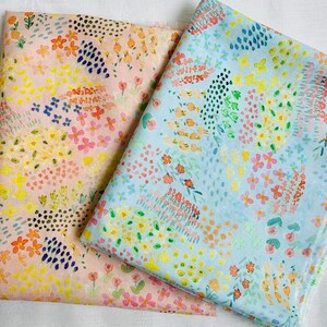 Könnte beinhalten: Zwei Stoffstücke mit einem Aquarell-Blumenmuster. Einer ist hellrosa, der andere hellblau. Beide haben ein sich wiederholendes Muster aus bunten Blumen, Punkten und Blättern in Rosa-, Gelb-, Orange- und Blautönen.