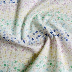 May include: White fabric with a repeating floral pattern in pastel colors. The design features small, four-petal flowers in shades of blue, green, yellow, and pink, with brown stems and leaves. The fabric is slightly wrinkled, suggesting a soft texture.