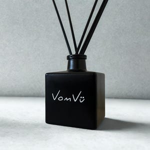 May include: A black square diffuser bottle with black reeds. The bottle has the white text "Vom Vü" on the front. The diffuser is a home fragrance product.