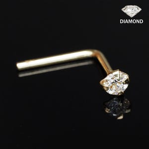 Könnte beinhalten: Ein goldfarbener Nasenstecker mit einem klaren Diamant-Edelstein. Der Stecker ist L-förmig mit einem kleinen, runden Diamanten am Ende. Das Wort "DIAMANT" steht in weißer Schrift über dem Stecker.