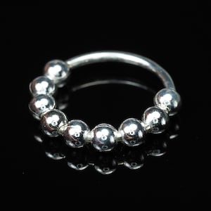 May include: A silver nose ring with a series of small, round beads. The beads are evenly spaced along the curved band, forming a continuous, circular design. The ring is set against a black background, accentuating its reflective surface.
