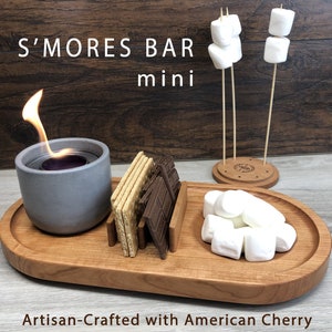 Könnte beinhalten: Ein Holztablett mit einem grauen Kerzenhalter aus Keramik, einem Stapel Graham-Cracker, einem Stapel Schokoladenquadraten und einem Haufen Marshmallows. Das Tablett ist mit "S'mores Bar mini" und "Artisan-Crafted with American Cherry" beschriftet.