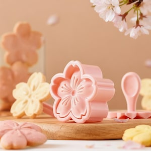 Peut inclure: Emporte-pièces roses en forme de fleur et biscuits sur une planche en bois. Les biscuits sont roses et jaunes. Un récipient en verre contient d'autres biscuits. Branche de fleurs de cerisier en arrière-plan.