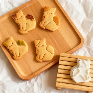 Puede incluir: Cuatro galletas con forma de gato con marcas marrones y verdes están dispuestas en una bandeja de madera clara. Un cortador de galletas blanco con forma de gato descansa sobre una pequeña plataforma de madera. Las galletas están sobre una superficie blanca texturizada.