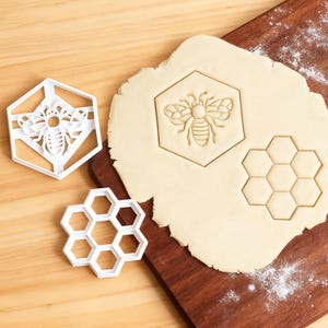 May include: White plastic bee and honeycomb-shaped cookie cutters next to a wooden cutting board with a cut-out bee and honeycomb shapes in dough. The bee cookie cutter is in a hexagon shape.