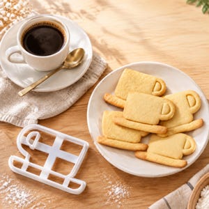 Op de afbeelding: Een witte plastic koekjesvorm in de vorm van een koffiemok met een oortje, geplaatst naast een wit bord met diverse gebakken, lichtbruine, mokvormige koekjes, een kop zwarte koffie en een goudkleurige lepel.