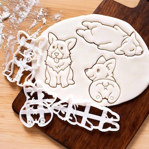 Könnte beinhalten: Ein Holzschneidebrett mit einem weißen Teigkreis mit Corgi-Hunde-Abdrücken. Weiße Corgi-förmige Ausstecher aus Kunststoff sind ebenfalls vorhanden. Der Teig hat Abdrücke eines sitzenden, liegenden und von hinten gesehenen Corgis.