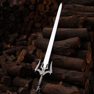 May include: A silver and white sword with an ornate handle and a long, straight blade. The sword is displayed against a backdrop of stacked brown logs, creating a rustic and dramatic contrast. The handle features intricate detailing.