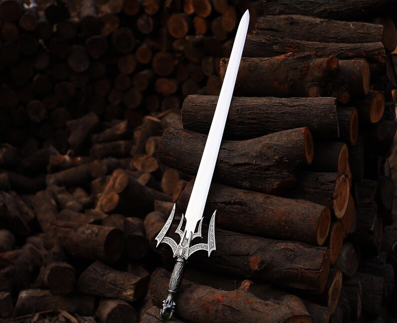 May include: A silver and white sword with an ornate handle and a long, straight blade. The sword is displayed against a backdrop of stacked brown logs, creating a rustic and dramatic contrast. The handle features intricate detailing.