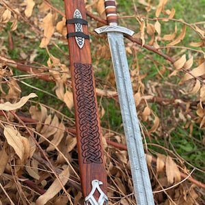 May include: A detailed image of a Viking sword with a patterned blade and wooden handle. The sword is accompanied by a wooden scabbard with intricate carvings and metal accents. The scabbard is brown with a black strap. The sword is resting on a bed of dry leaves and twigs.