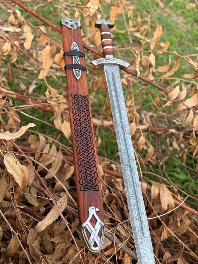 May include: A detailed image of a Viking sword with a patterned blade and wooden handle. The sword is accompanied by a wooden scabbard with intricate carvings and metal accents. The scabbard is brown with a black strap. The sword is resting on a bed of dry leaves and twigs.