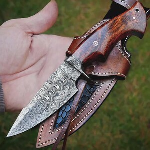 May include: A handcrafted hunting knife with a patterned steel blade and a dark wood handle. The knife is housed in a brown leather sheath with decorative stitching. The blade has a unique, swirling pattern.