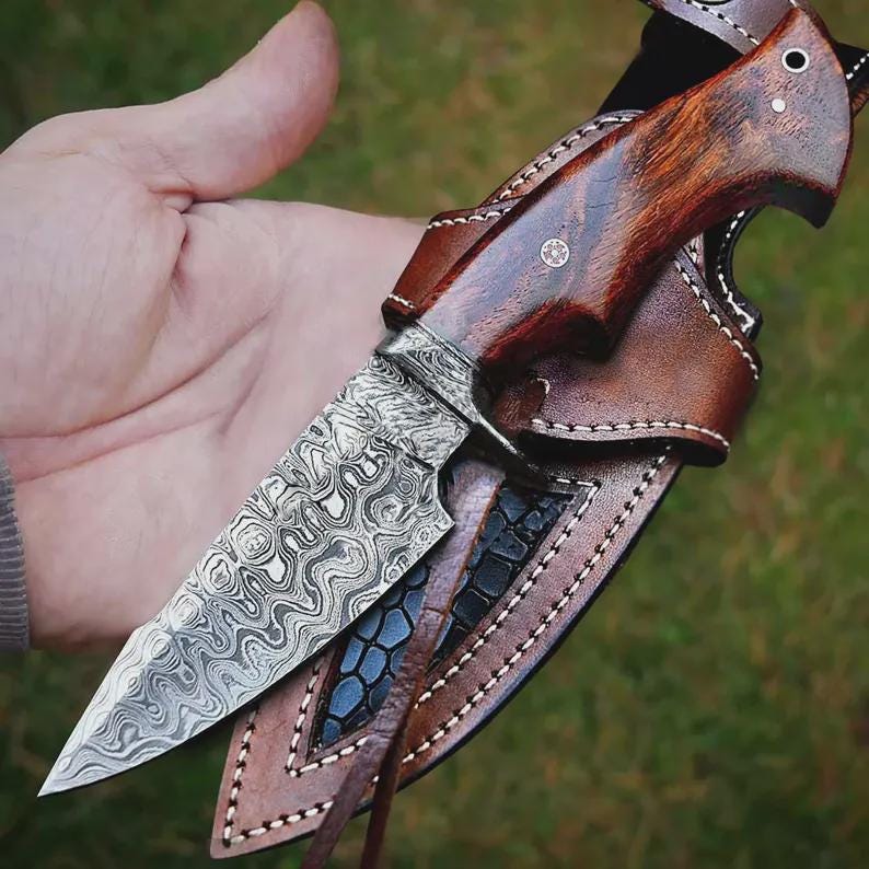 May include: A handcrafted hunting knife with a patterned steel blade and a dark wood handle. The knife is housed in a brown leather sheath with decorative stitching. The blade has a unique, swirling pattern.