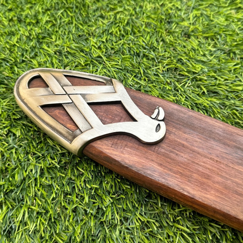 May include: Close-up of a wooden scabbard with a decorative metal fitting. The metal piece has a Celtic knot design and a curved, stylized shape. The wood is a rich brown color, and the metal has a bronze finish.