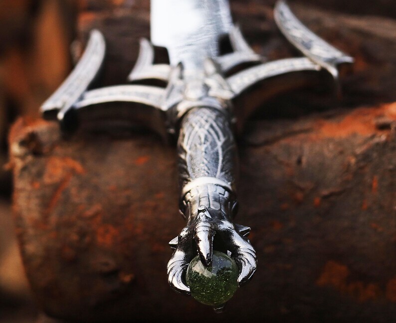 May include: Close-up of a decorative sword with a silver blade and ornate handle. The handle features intricate carvings and a claw-like design holding a green gemstone. The sword is set against a blurred, textured background.