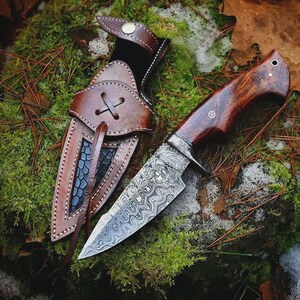 May include: A handcrafted hunting knife with a patterned steel blade and a dark wooden handle. The knife is paired with a brown leather sheath, featuring decorative stitching and a cross-stitched closure. The knife and sheath are set against a backdrop of moss and foliage.