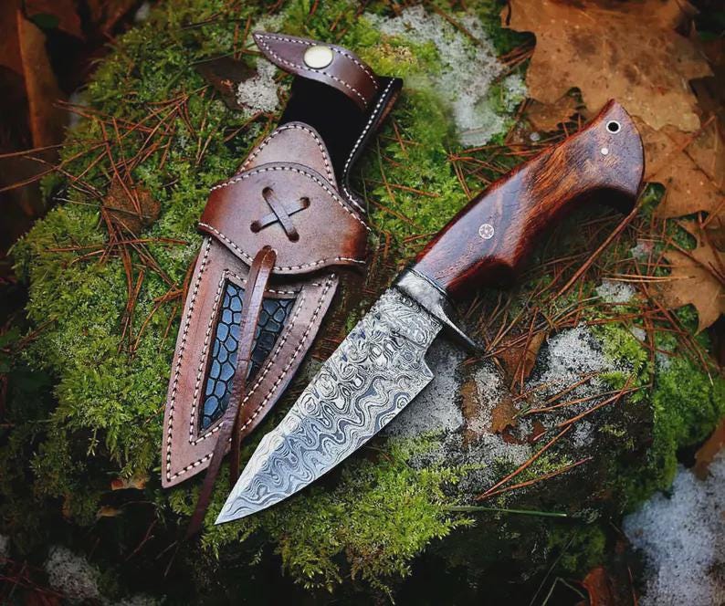 May include: A handcrafted hunting knife with a patterned steel blade and a dark wooden handle. The knife is paired with a brown leather sheath, featuring decorative stitching and a cross-stitched closure. The knife and sheath are set against a backdrop of moss and foliage.
