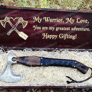 May include: A Viking axe with a silver head and a dark wooden handle, presented in a wooden box. The box is engraved with the words "My Warrior, My Love" and "Happy Gifting!"