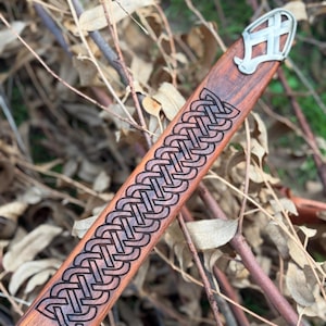 May include: A wooden sword scabbard with a decorative Celtic knot design carved into the wood. The scabbard has a metal end piece with a stylized design. The wood has a reddish-brown color.