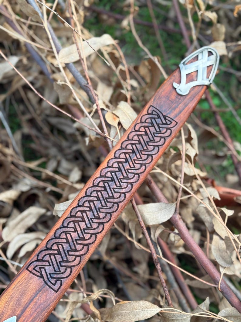 May include: A wooden sword scabbard with a decorative Celtic knot design carved into the wood. The scabbard has a metal end piece with a stylized design. The wood has a reddish-brown color.