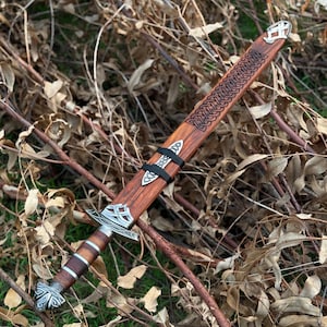 May include: A wooden sword with a brown sheath and intricate silver-colored metal detailing. The sheath features a carved design and is secured with a black strap. The sword's handle is wrapped in brown leather.