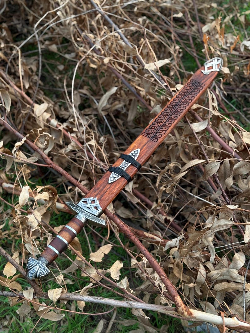 May include: A wooden sword with a brown sheath and intricate silver-colored metal detailing. The sheath features a carved design and is secured with a black strap. The sword's handle is wrapped in brown leather.