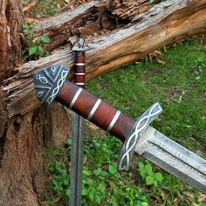 May include: Two swords with ornate silver and wood handles rest on a bed of green grass. The blades have a detailed pattern. One sword is in focus, while the other is slightly blurred in the background. The swords are near a fallen tree.