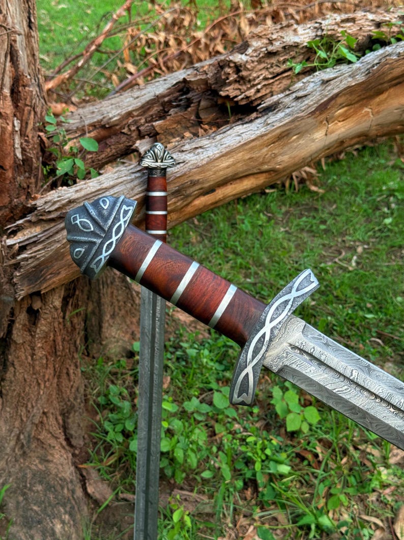 May include: Two swords with ornate silver and wood handles rest on a bed of green grass. The blades have a detailed pattern. One sword is in focus, while the other is slightly blurred in the background. The swords are near a fallen tree.
