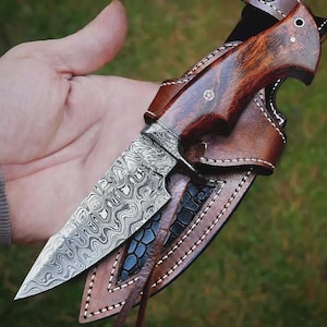 May include: A handcrafted hunting knife with a patterned steel blade and a dark wood handle. The knife is housed in a brown leather sheath with decorative stitching. The blade has a unique, swirling pattern.