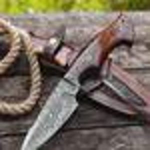 May include: A handcrafted hunting knife with a patterned steel blade and a dark wood handle. The knife rests on a leather sheath, with a rope in the background. The blade measures approximately 8 inches.