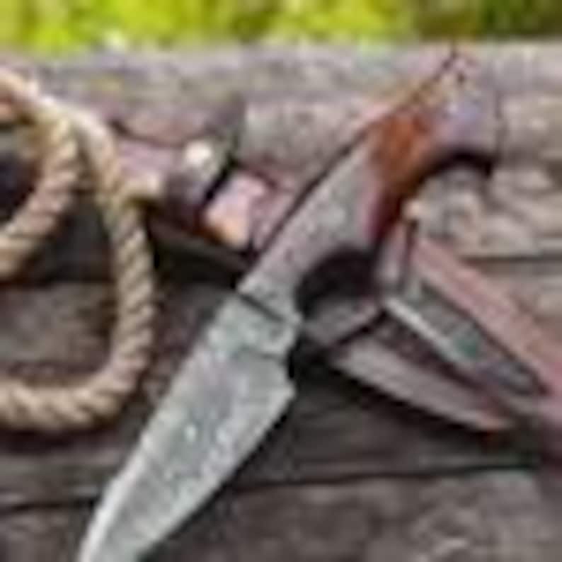 May include: A handcrafted hunting knife with a patterned steel blade and a dark wood handle. The knife rests on a leather sheath, with a rope in the background. The blade measures approximately 8 inches.