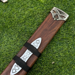 May include: A wooden sword scabbard with silver-colored metal accents. The scabbard is brown with a decorative metal end cap and two bands. The bands and end cap feature Celtic knot designs. The scabbard is lying on a bed of green grass.