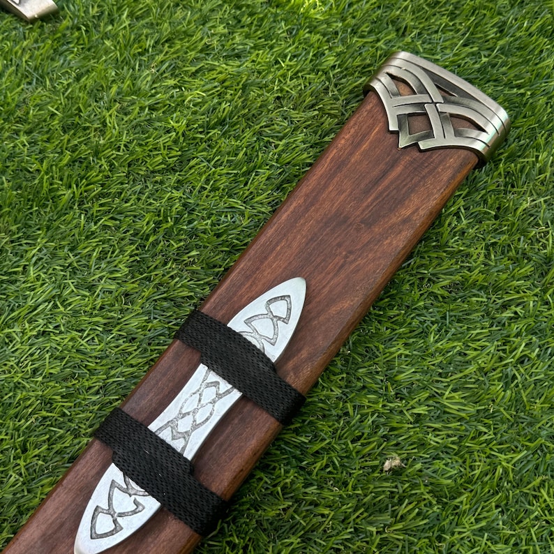 May include: A wooden sword scabbard with silver-colored metal accents. The scabbard is brown with a decorative metal end cap and two bands. The bands and end cap feature Celtic knot designs. The scabbard is lying on a bed of green grass.