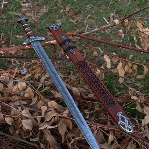 May include: A detailed image of a Viking sword with a silver blade and a wooden handle. The sword is displayed next to its wooden sheath, which features intricate Celtic knotwork and metal accents. The sword and sheath are resting on a bed of twigs and leaves.
